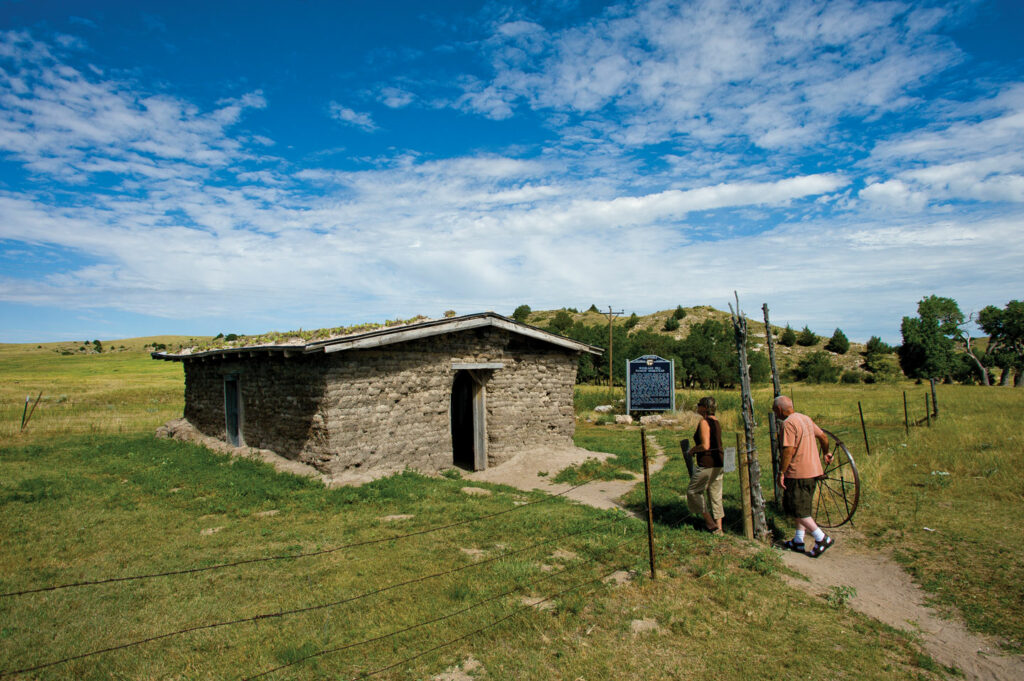 A man and woman walk up to a sod house in under vivid blue skies.