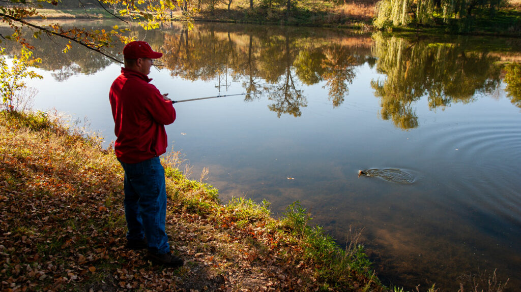 a man fishes from the bank of a lake surrounded by fall foliage