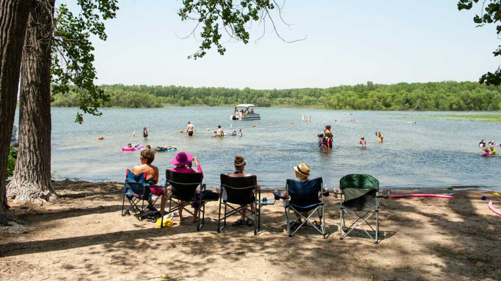 Four people sit in camp chairs watching visitors swim in the lake. A pontoon boat is seen in the distance.