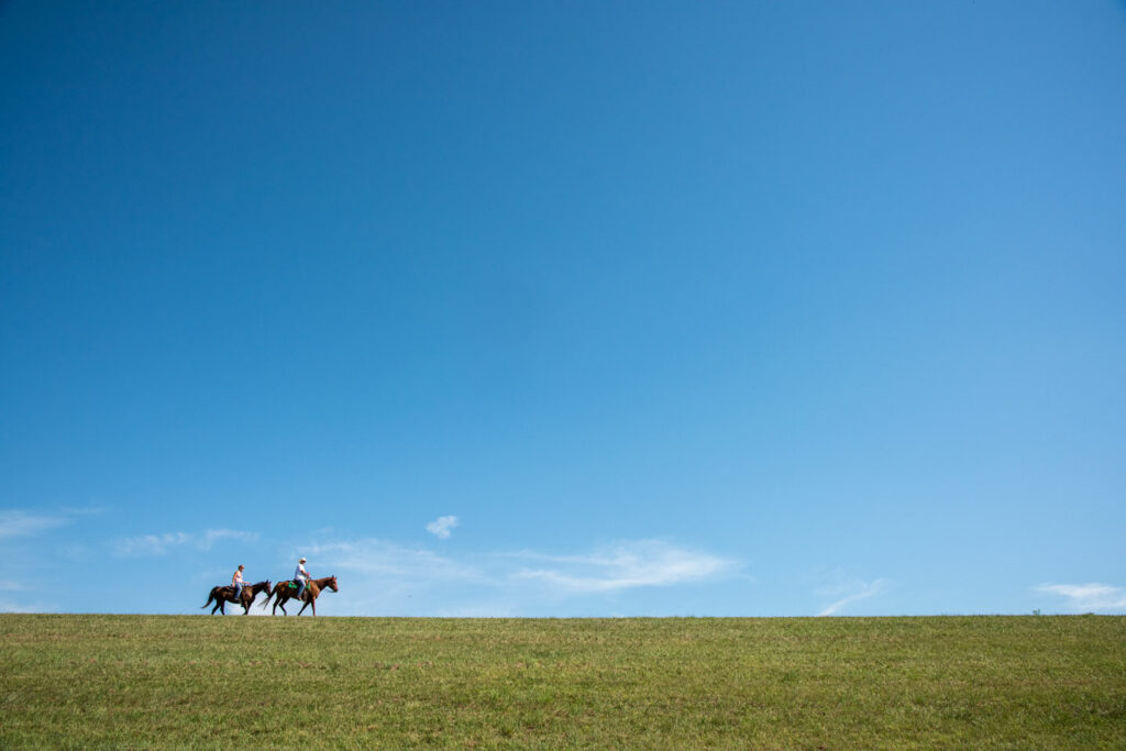 Two people ride horses on the green landscape under a clear blue sky.