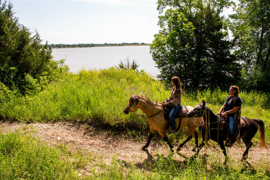 Two women ride horses on a trail by the lake