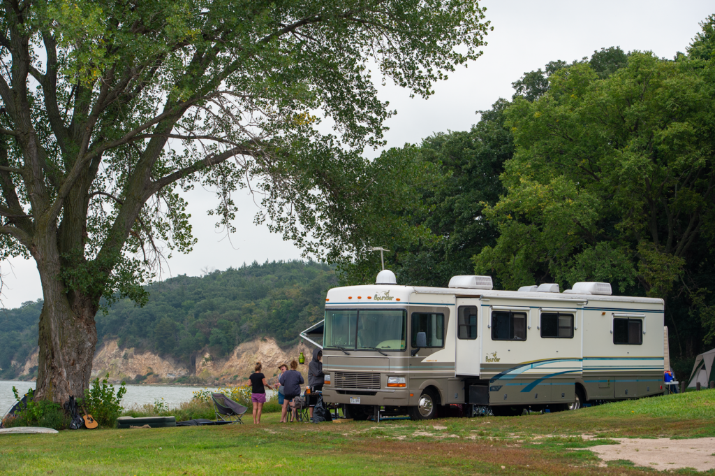 An RV camper is parked next to a tree line along the shore and a tree-covered bluff