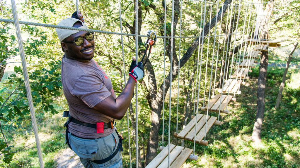 a young man smiles at the camera as he navigates the rope course suspended from the trees