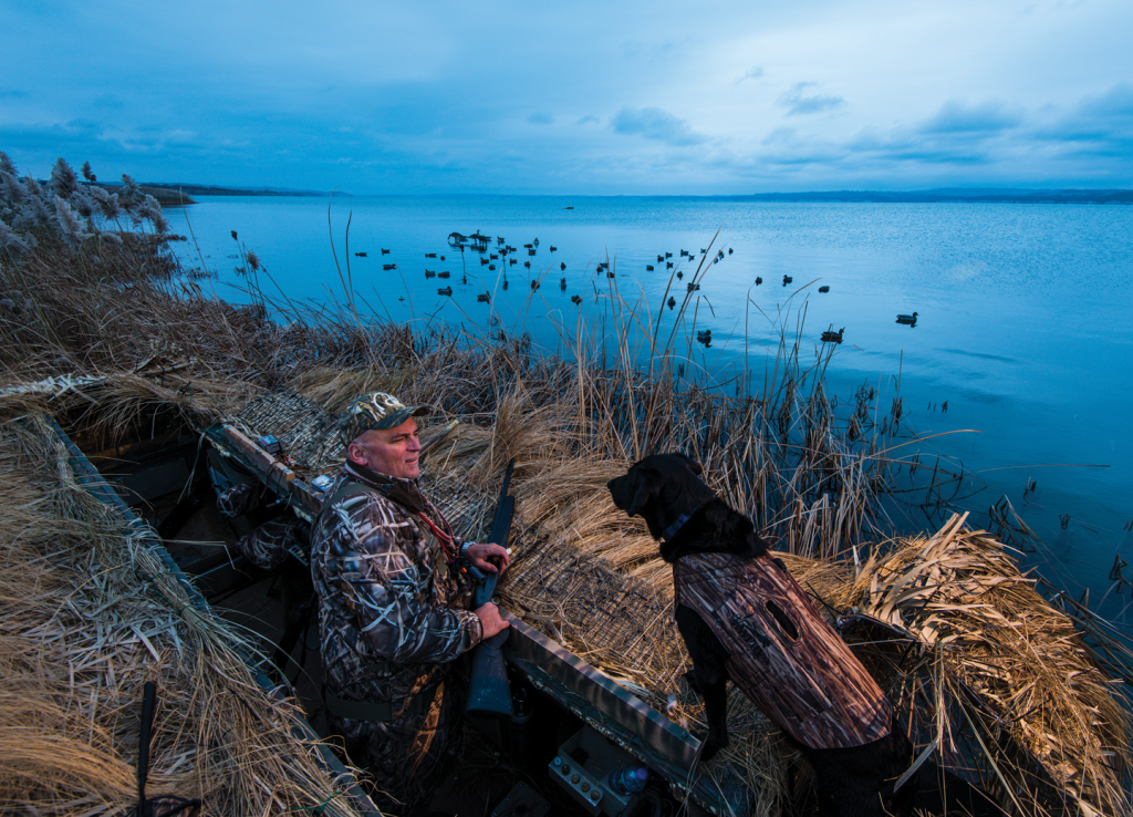 A man and his black lab stand in a duck blind