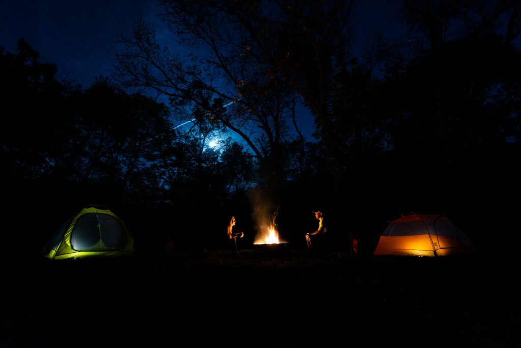 a young girl and a man sit around a campfire near their tents at night with the moon shining
