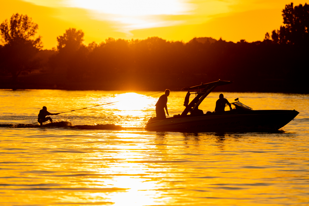 A wakeboarder and a boat full of people are silhouetted against a yellow sky