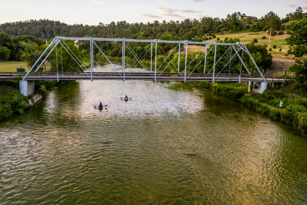 Two kayaks are directly under a steel bridge that goes across a river