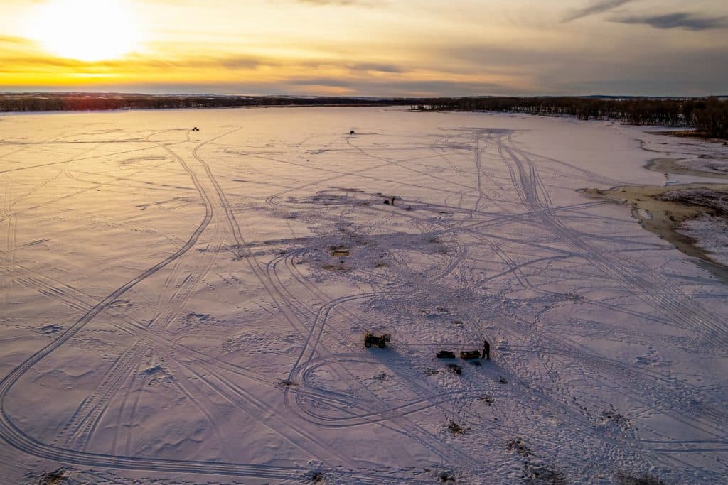 Aerial view above a frozen lake covered in ice fishing tracks as the sun beams on the horizon