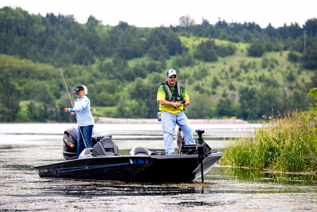 a man and woman stand on a bass boat fishing near the lake's edge
