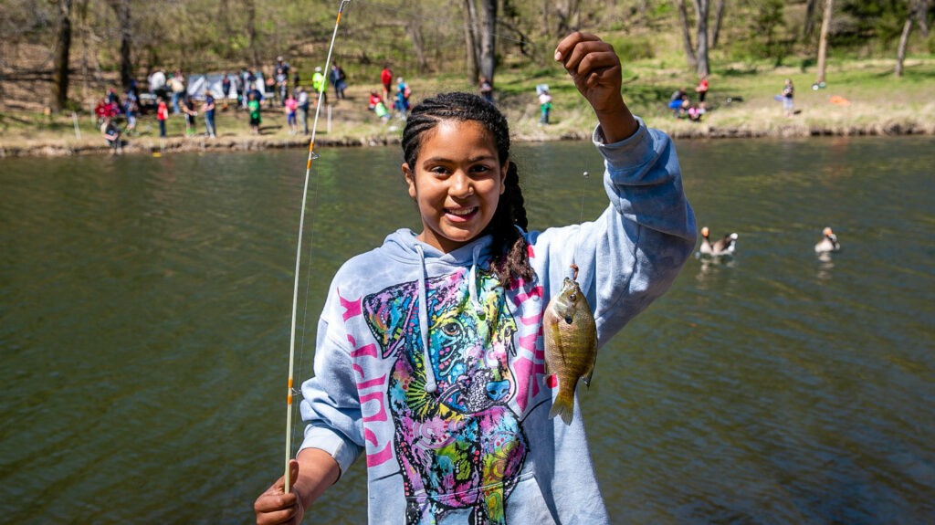 a young girl stands on the bank of the lake, holding a fish that she caught on a line