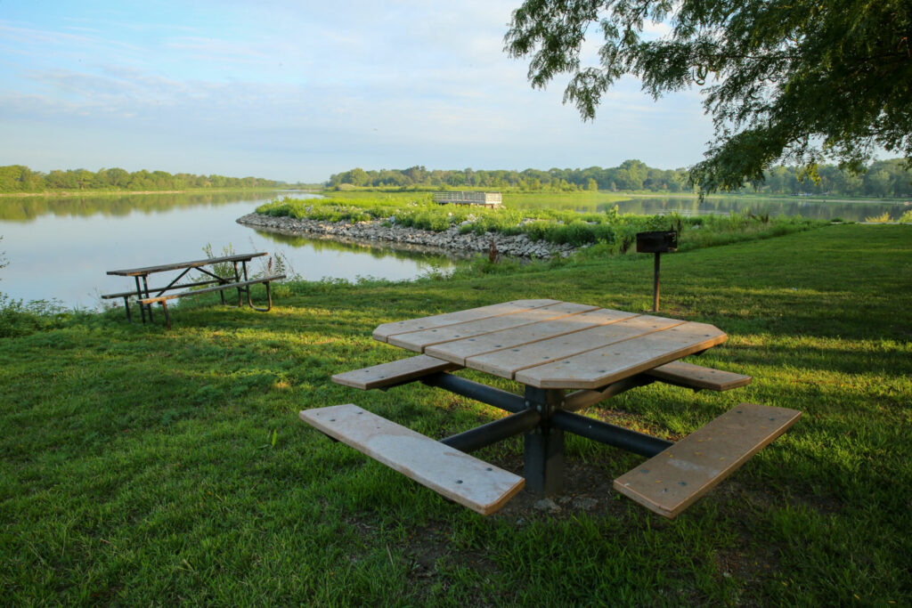 As square picnic table is in the foreground with a lake in the background.