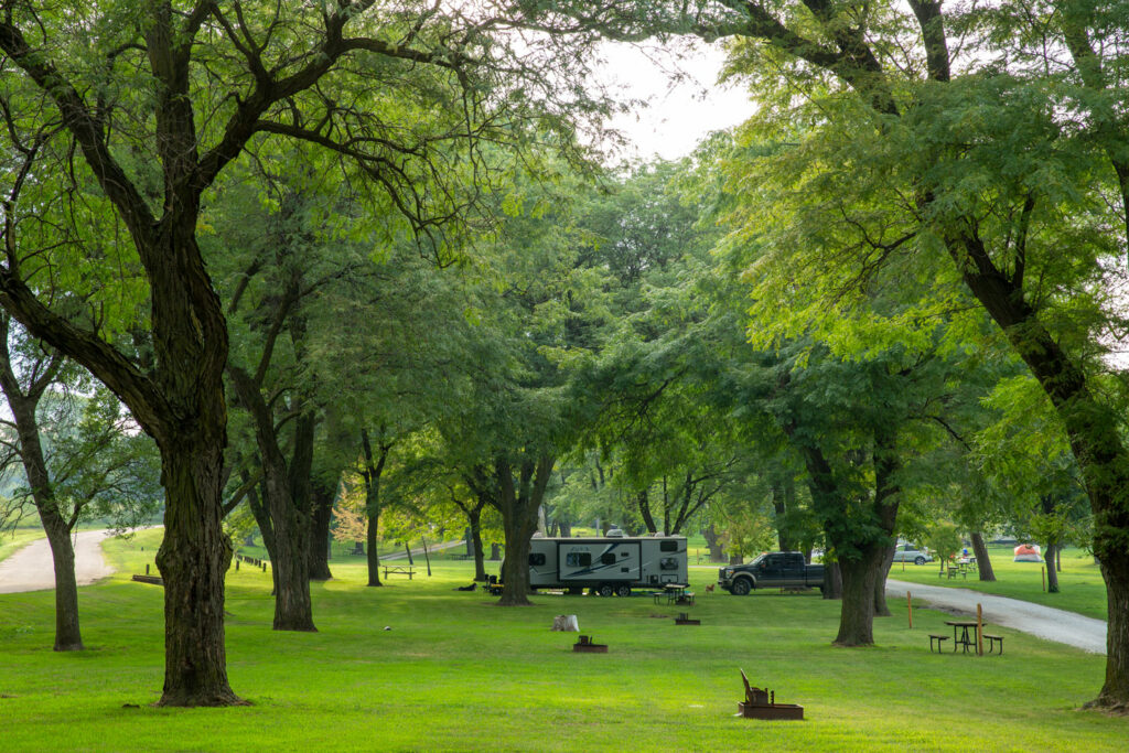 An RV camper is framed by tall deciduous trees in a campground at Memphis State Recreation Area.