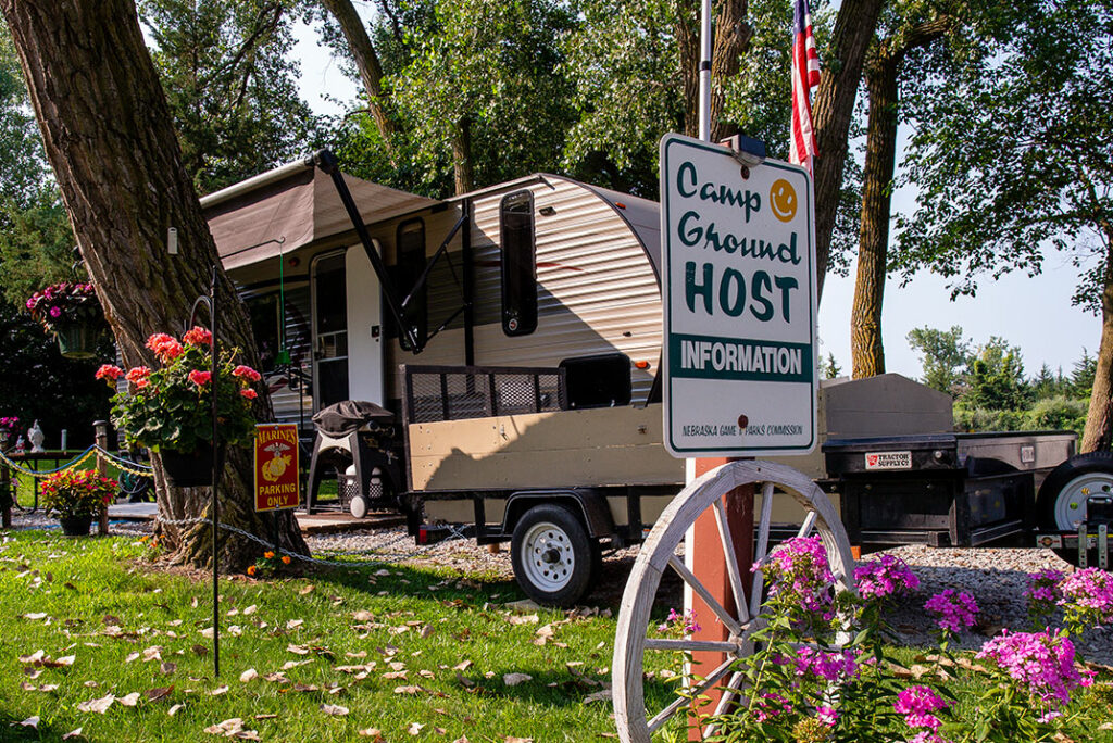 A sign reading "Campground Host" is shown in front of an occupied RV site with blooming potted flowers around.
