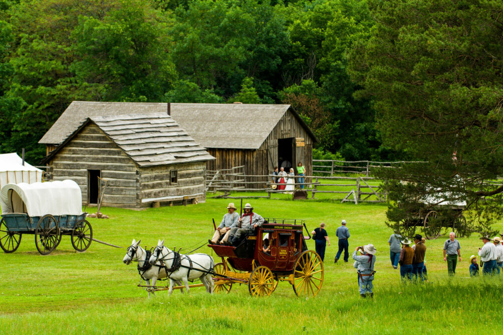 Two white horses pull a red stagecoach across the grass; old wooden buildings are in the background, and people mill about Rock Creek Station State Historical Park.