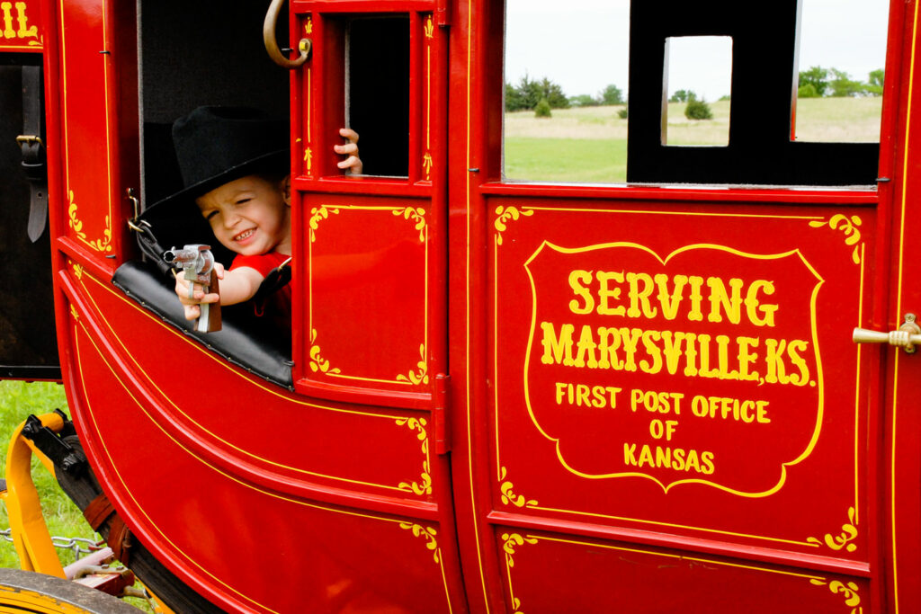 A little boy rides in a red stagecoach, pretending to shoot a toy pistol out the window.