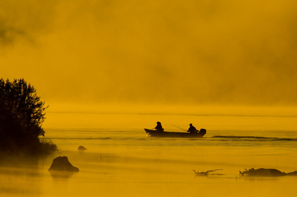 A boat with two anglers in it are silhouetted against a yellow sky.
