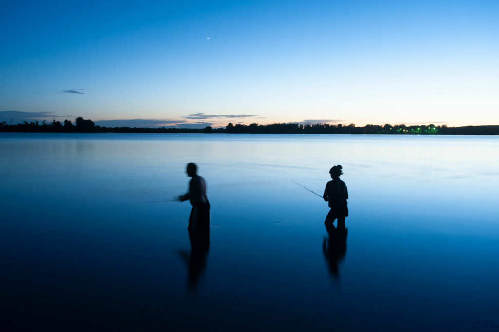 A man and woman are silhouetted standing knee deep in water fishing.