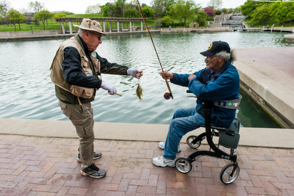 A man helps another in a wheelchair unhook a bluegill from his hook while fishing.