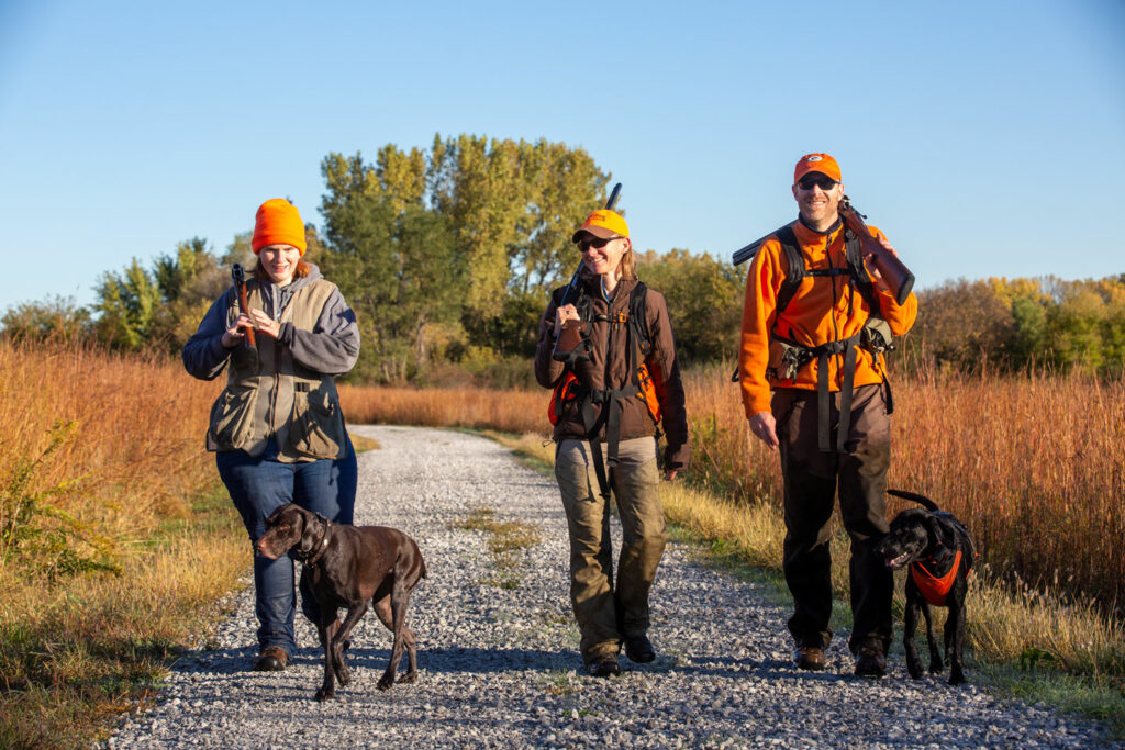 Two women, a man, and two pointer dogs walk down a gravel driveway.