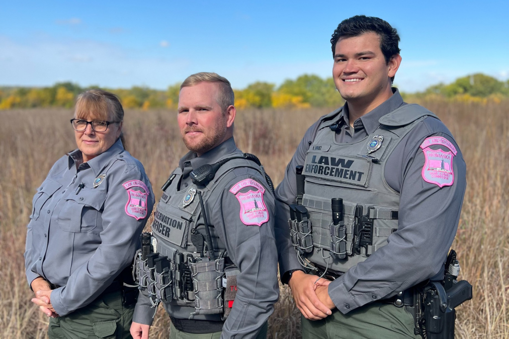 Three Nebraska Game and Parks Conservation Officers standing in a field wearing uniforms with breast cancer awareness patches