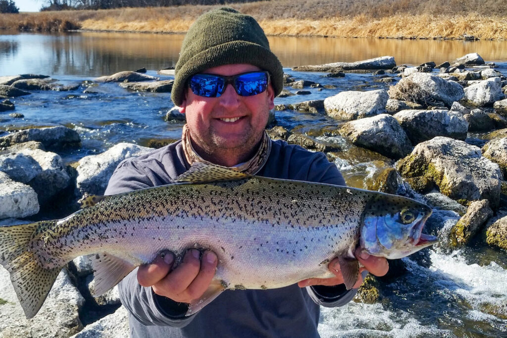 a man holds a large trout in front of a stream