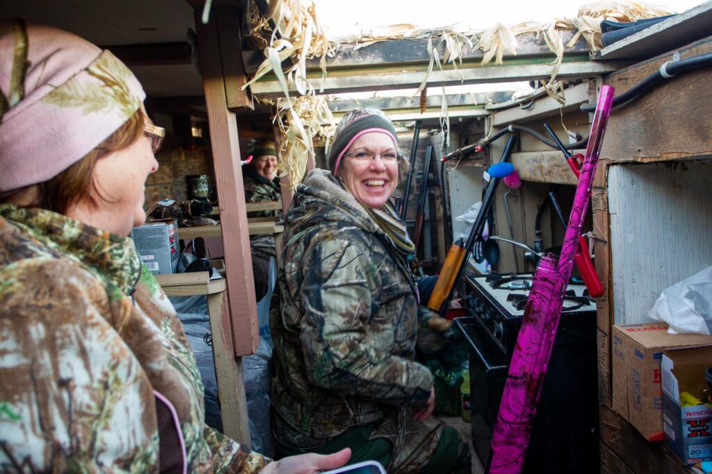 three smiling women in hunting gear in a waterfowl blind