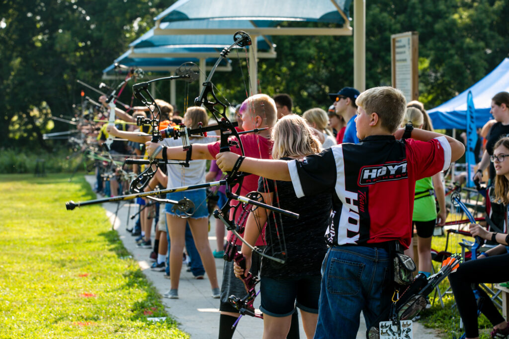 A group of kids line up with archery equipment while competing in an outdoor archery tournament at NOEC