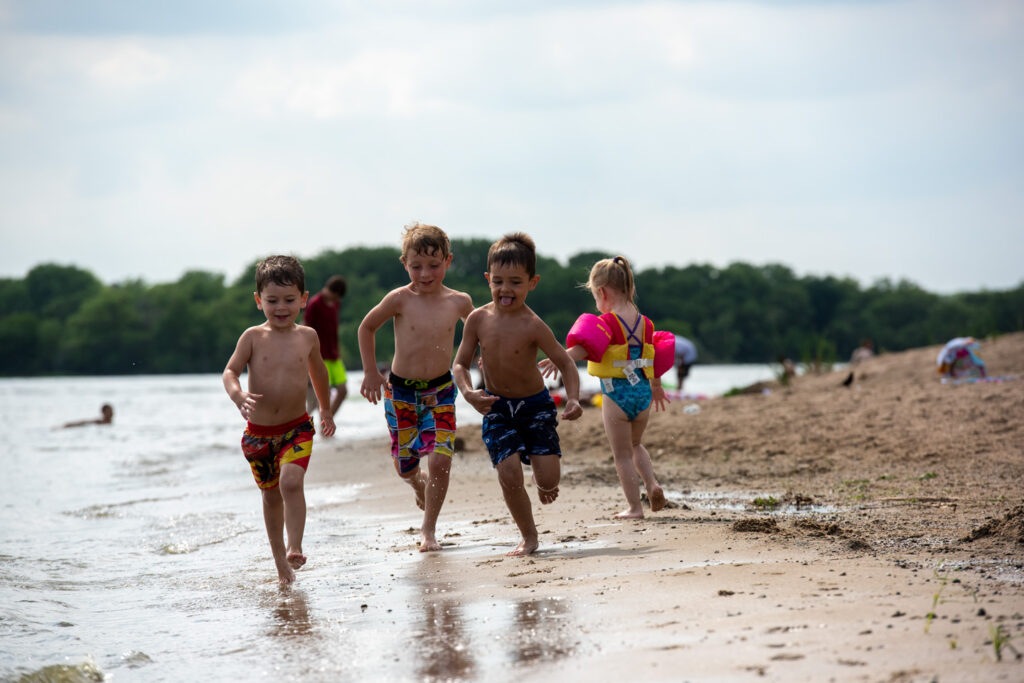 Three boys run down the beach at the edge of the water.
