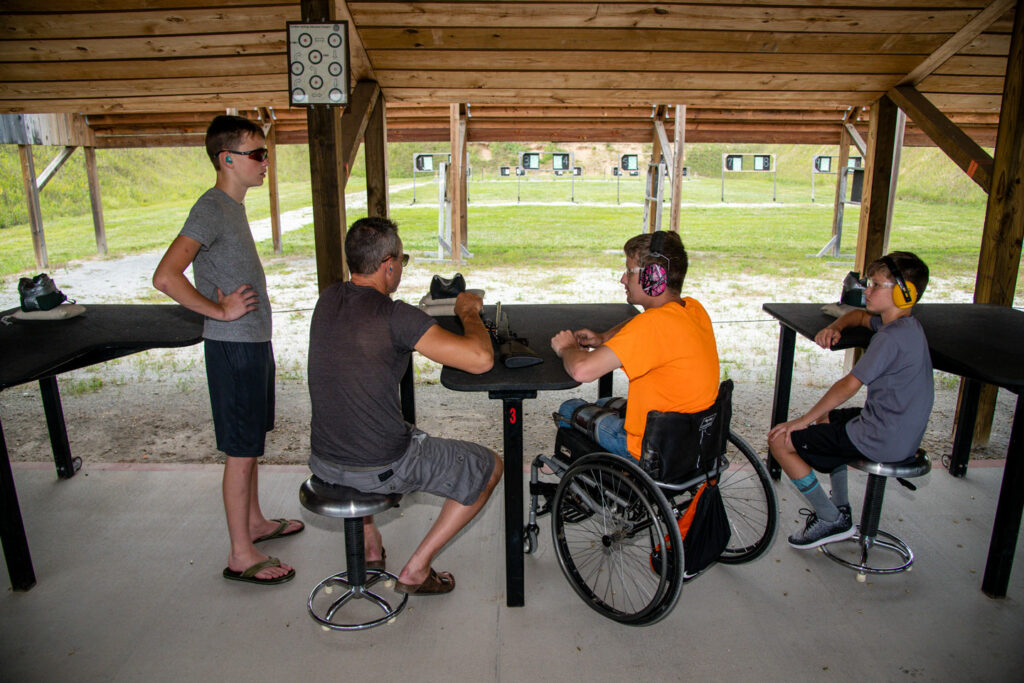 A young man in a wheelchair is coached to shoot a rimfire rifle.