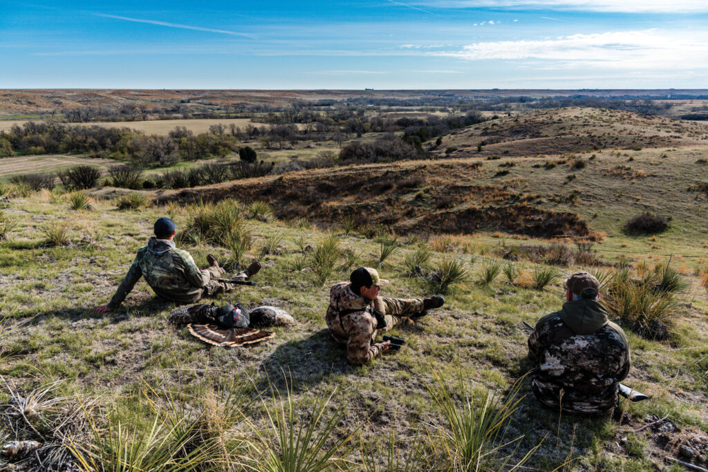 Three men sit back on a crest looking for turkeys. 