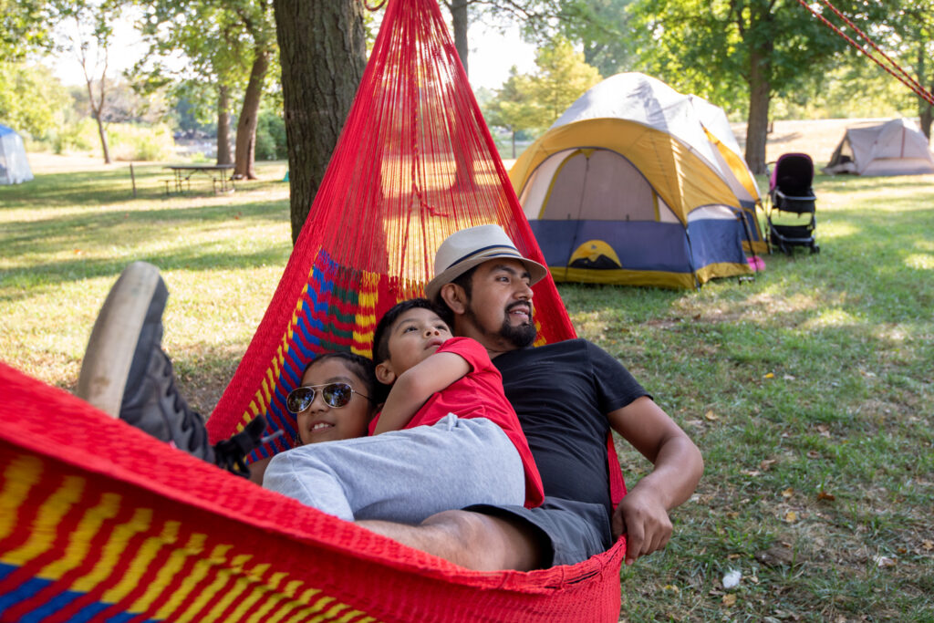 A man and his two children lay in a hammock in front of a tent
