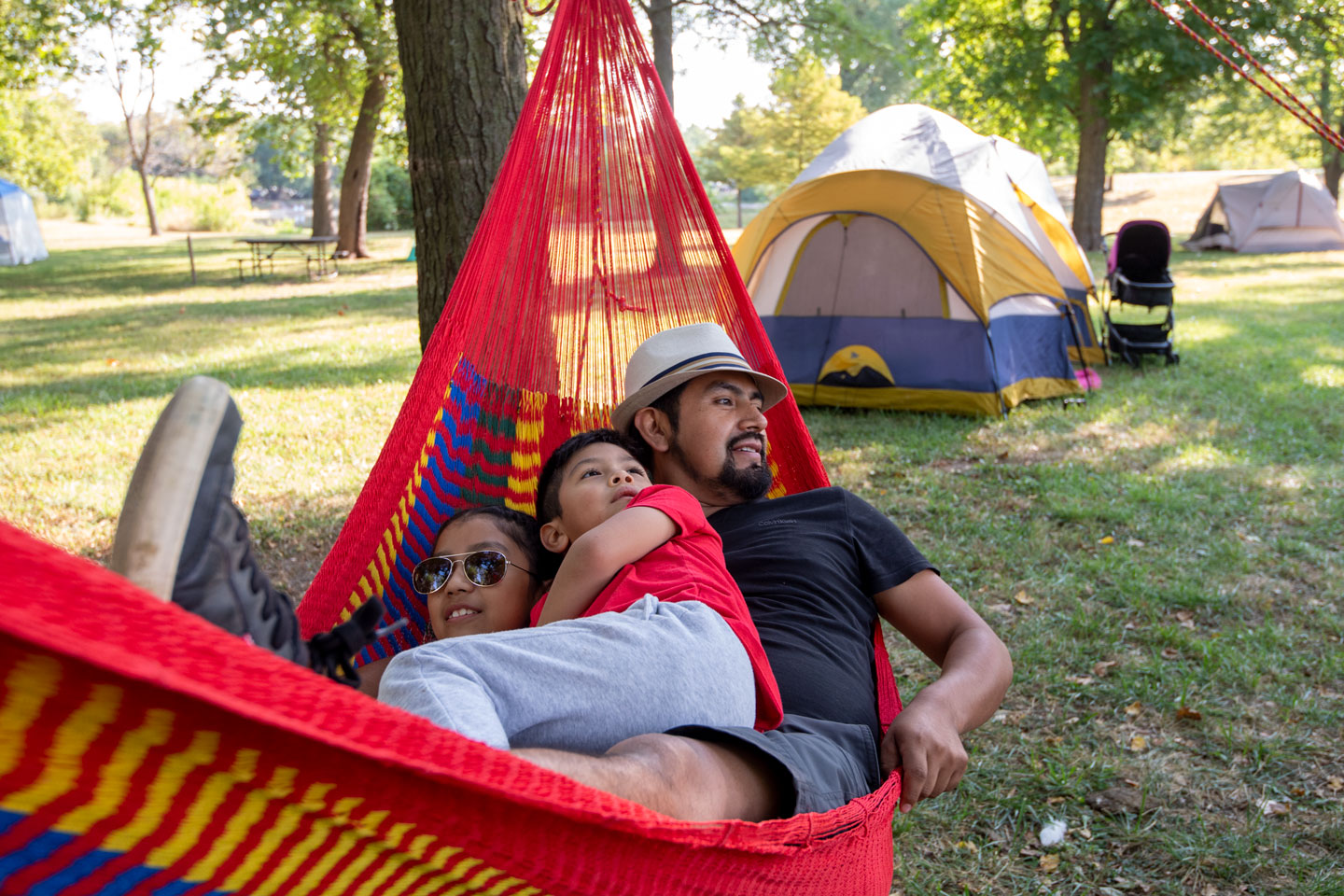 A man and two children lay in a red hammock at a campground.