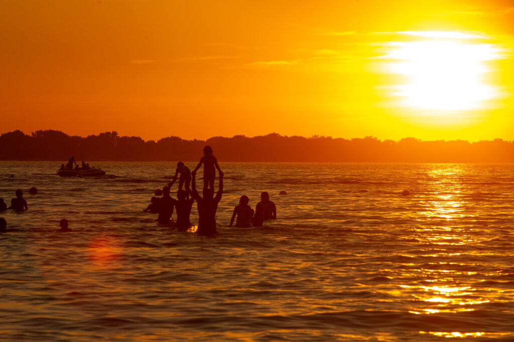 People standing on others' shoulders in the water are silhouetted by a setting sun.