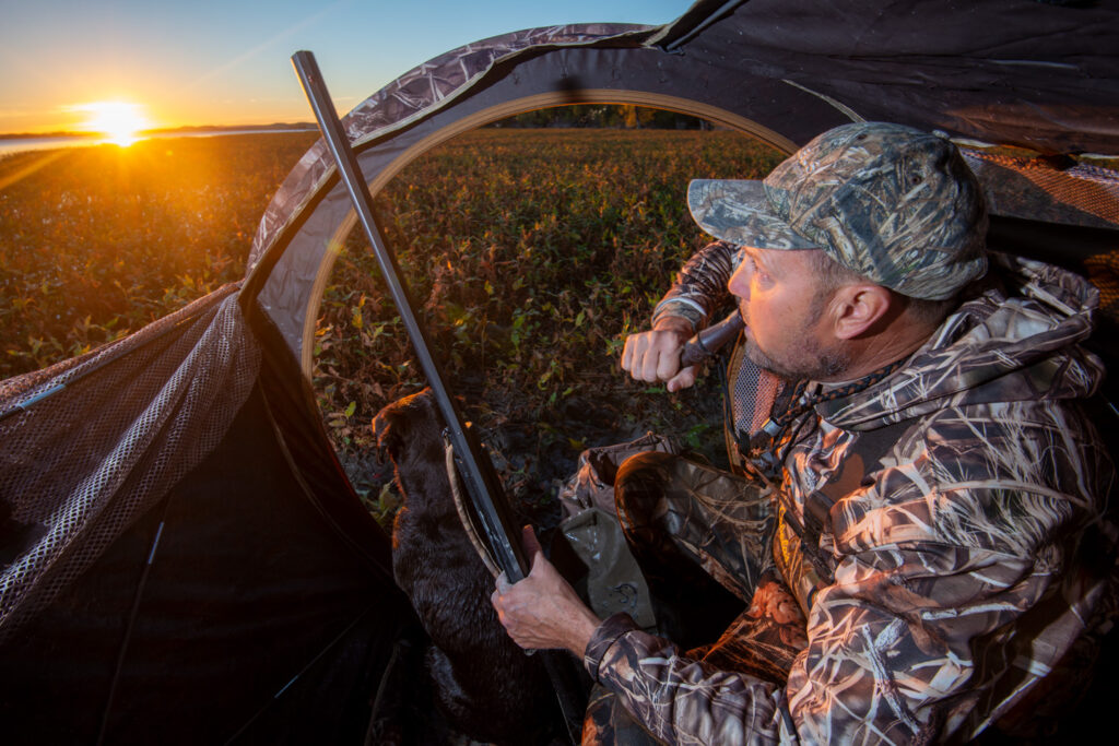 A mam uses a duck call as the sun peaks over the horizon at sunrise.