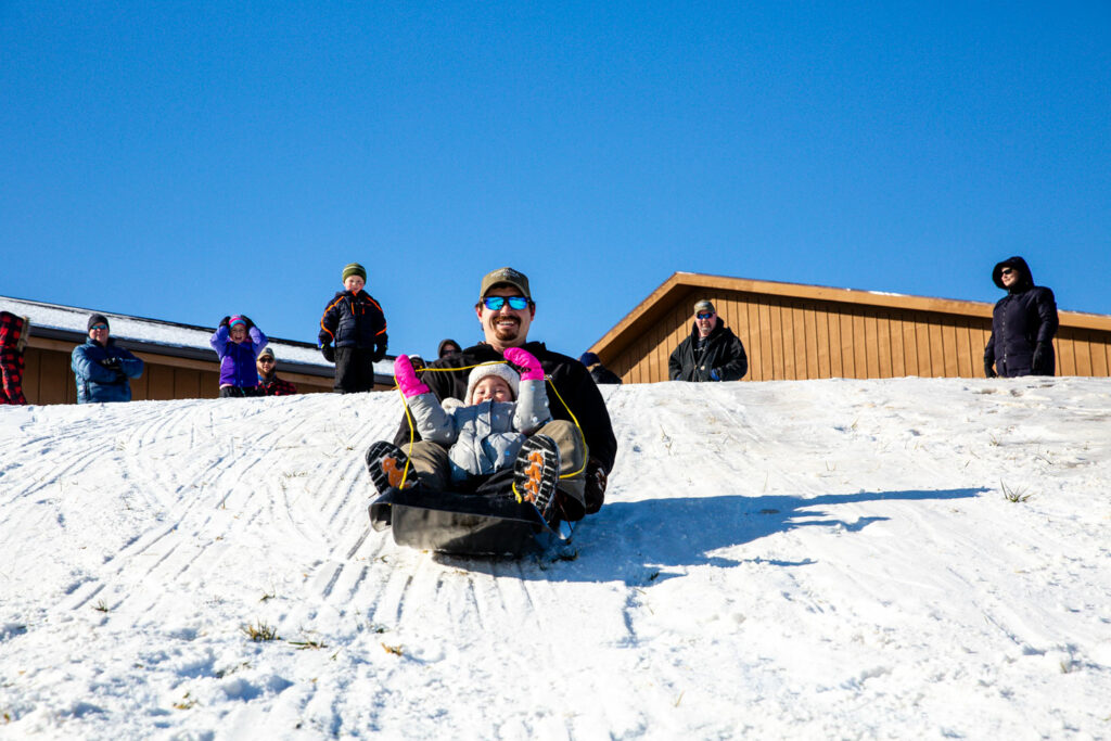 a father and daughter share a sled as they laugh and slide down the snowy hill