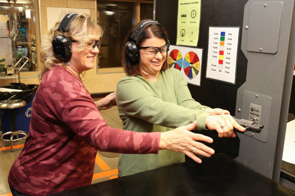 A woman coaches another how to shoot a pistol.