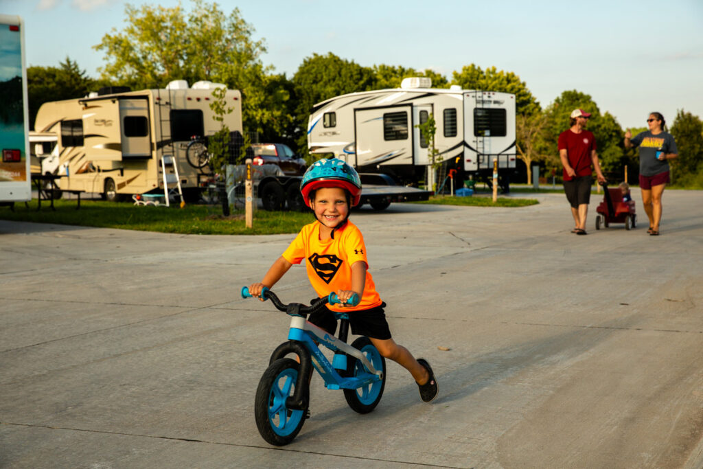 a young boy on a bike smiles at the camera while parents walk behind him in an RV campground