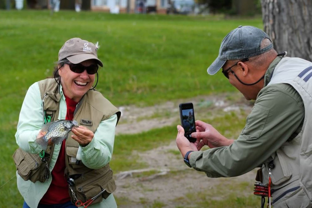 A smiling woman holds a panfish as her friend takes a cell phone photo of her.