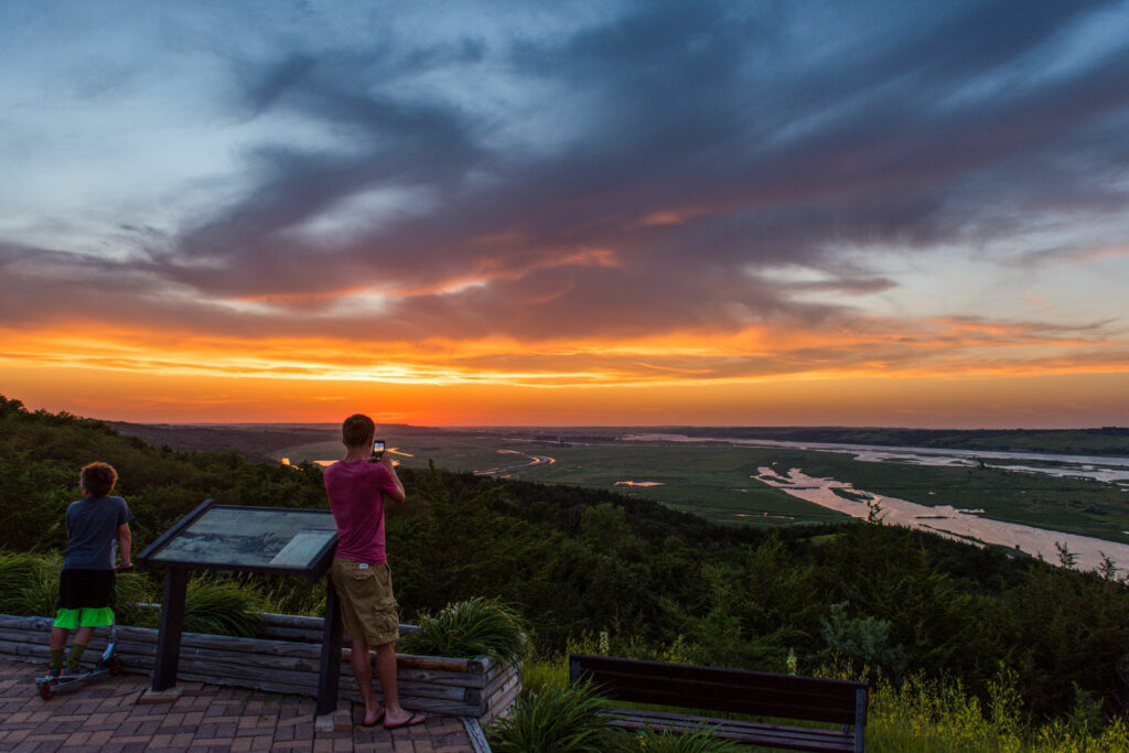 Visitors watch the sun set over the Missouri River bottom from Niobrara State Park in Knox County. Much of the bottomland and backwaters in the photo are part of Niobrara Confluence WMA. Fowler, June 9, 2017. Copyright NEBRASKAland Magazine, Nebraska Game and Parks Commission.