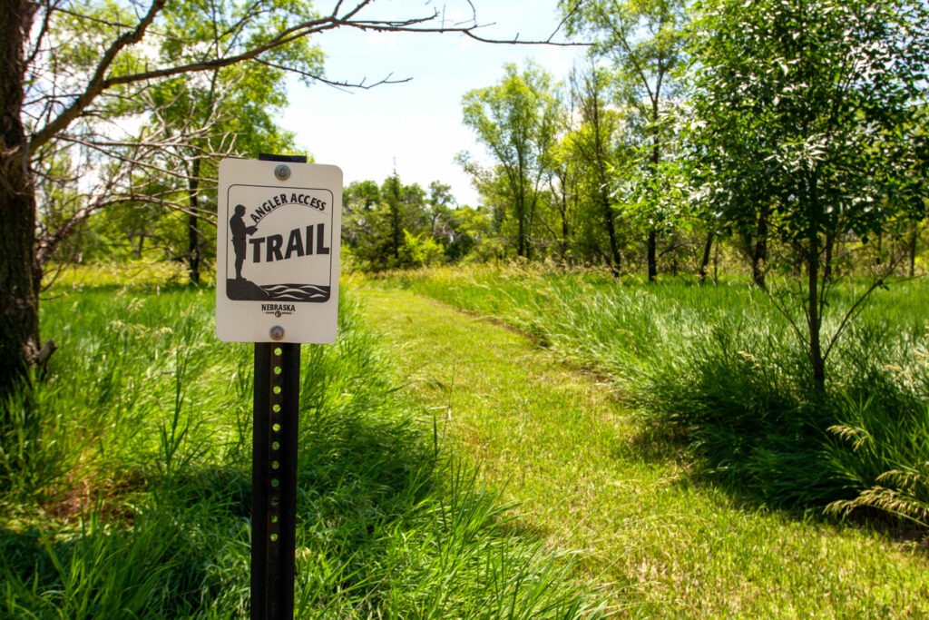 An angler access trail sign stands in the foreground of a grassy trail at a park.