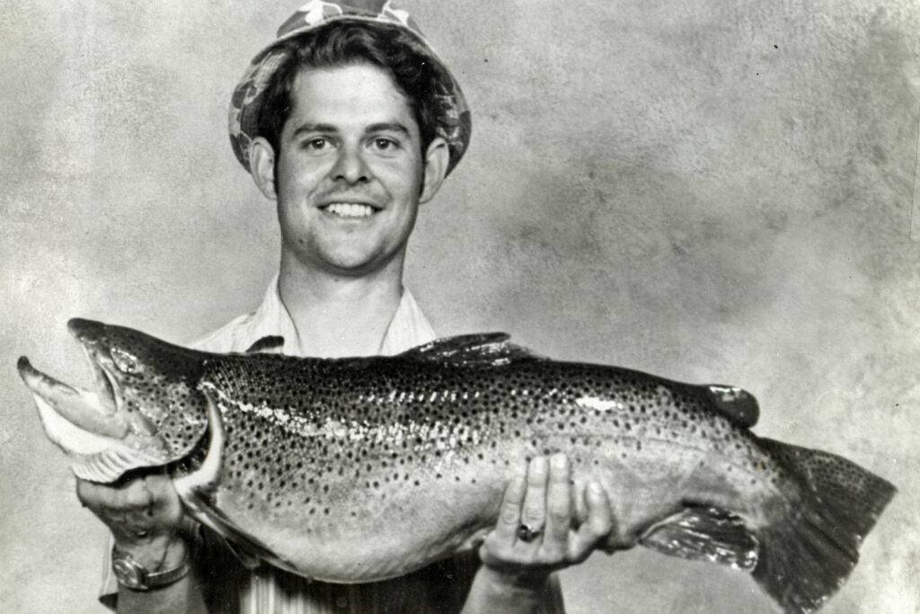 A man poses with his Master Angler Award state record-sized brown trout fish in a vintage black and white photo taken in 1973 in Nebraska