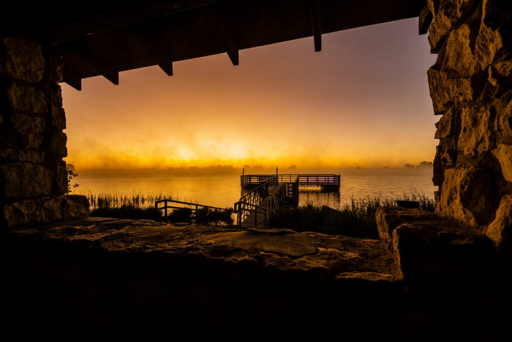 View if a sunrise through a stone shelter and fishing pier at Walgren Lake as mist billows from the water.