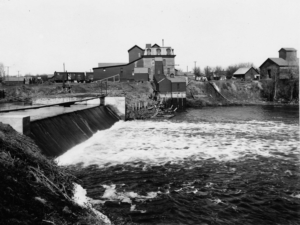 A black and white photo of an old mill taken somewhere between 1904-1909 with a river in the foreground.