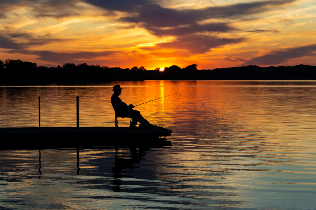 Silhouette of a man fishing on a dock at sunset on a lake.