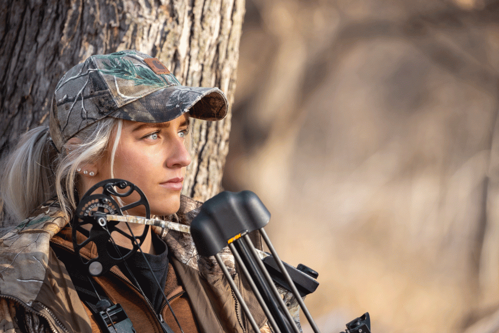 A woman sits in front of a tree with her archery bow