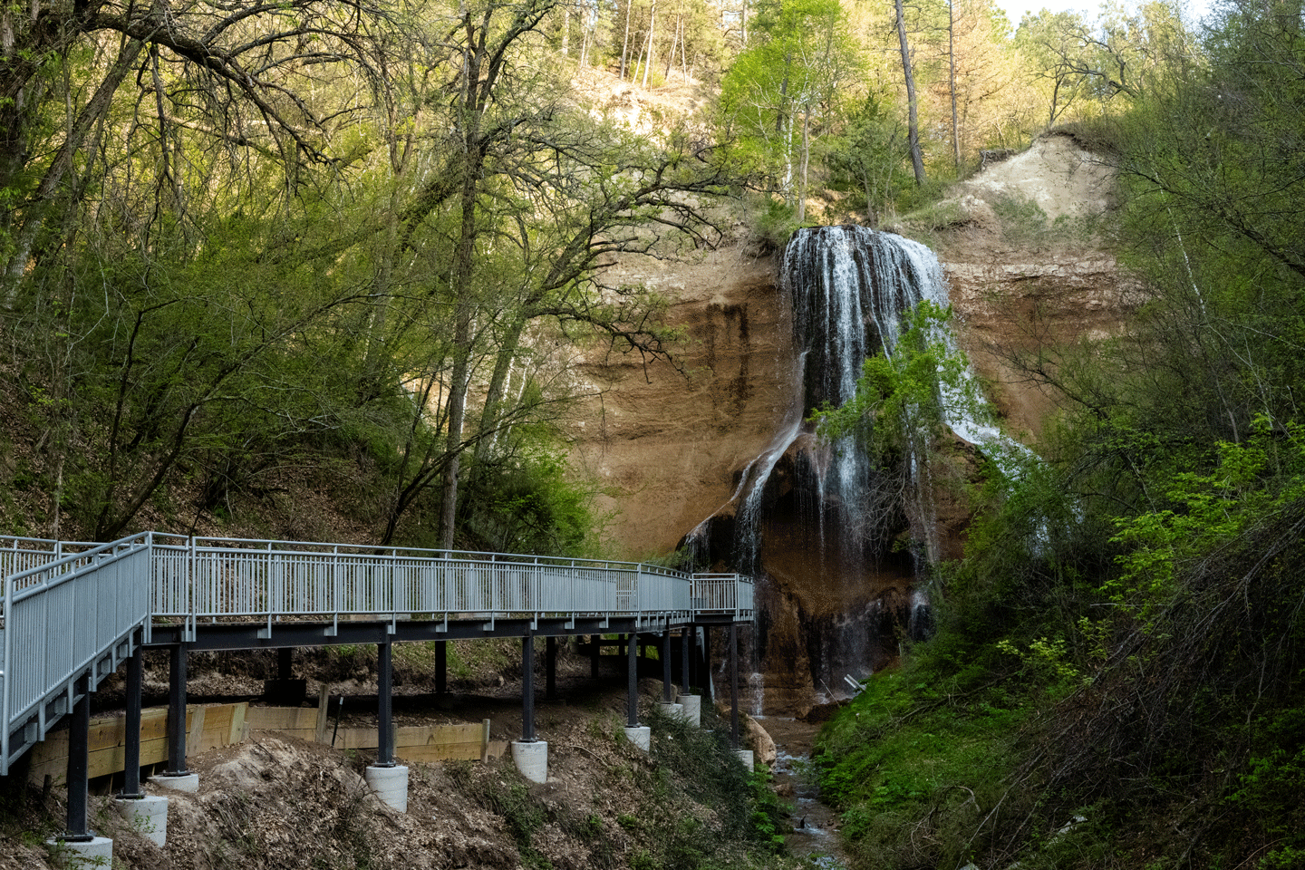 a metal and composite boardwalk is suspended above a creek below a waterfalk