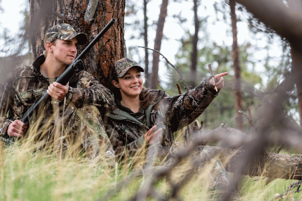 A man and woman sit on the ground in grassy cover while turkey hunting