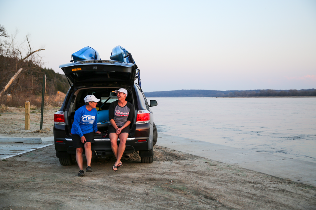 two women sit on the tail of the SUV, their kayaks on top of the vehicle