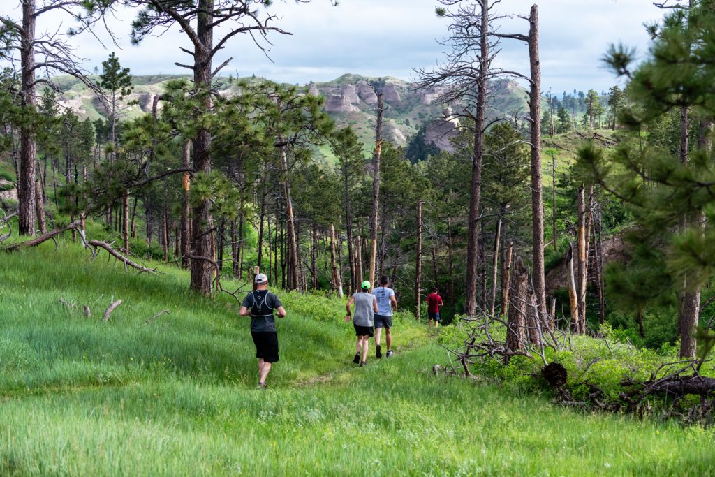 Runners run down a hiking trail in a forested area of Nebraska's Pine Ridge at Chadron State Park with butte formations in the background.