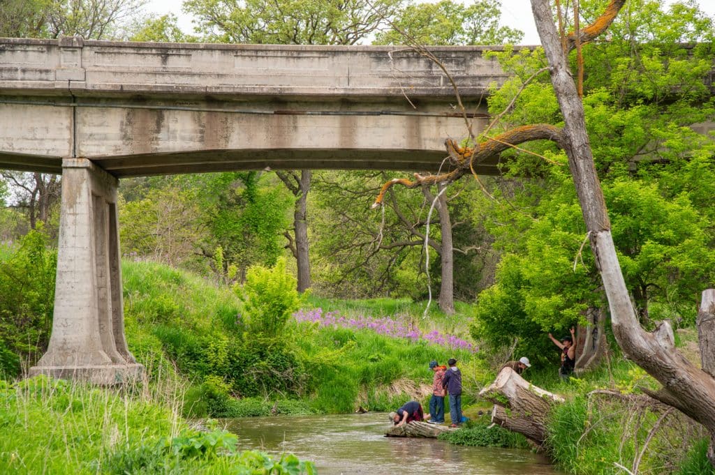 A group of school boys plays at the edge of a winding creek in a wooded area near a bridge during springtime.